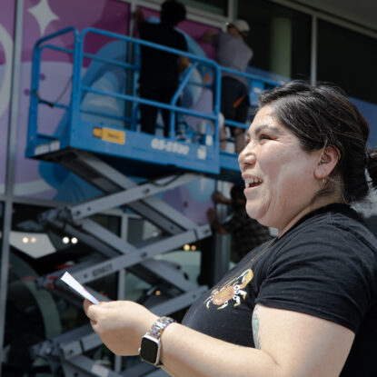 Artist Angelina Villalobos looking to the side as her mural is being installed in the background.