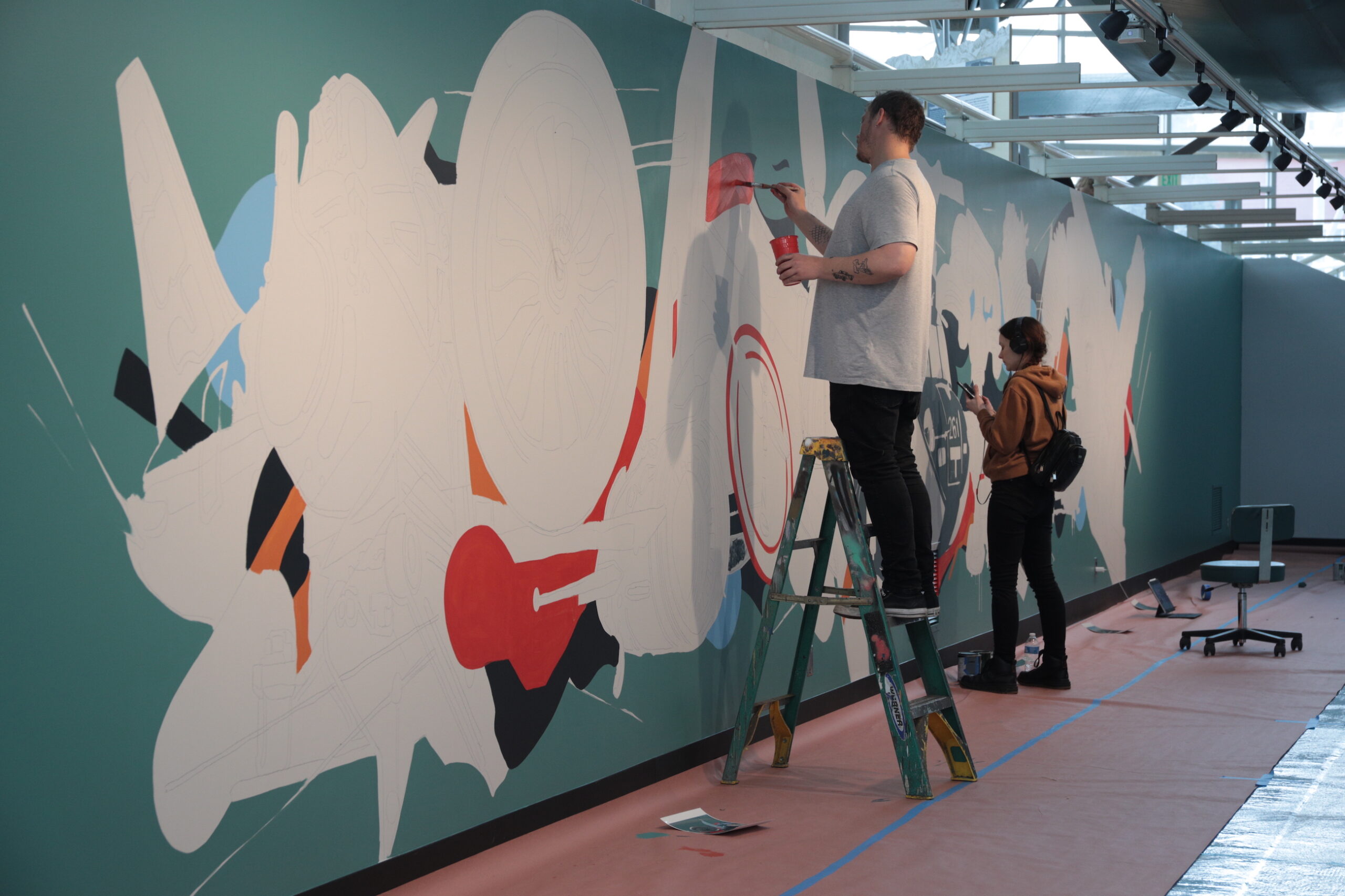 A partially finished mural is being worked on by two painters, with one standing on a ladder.