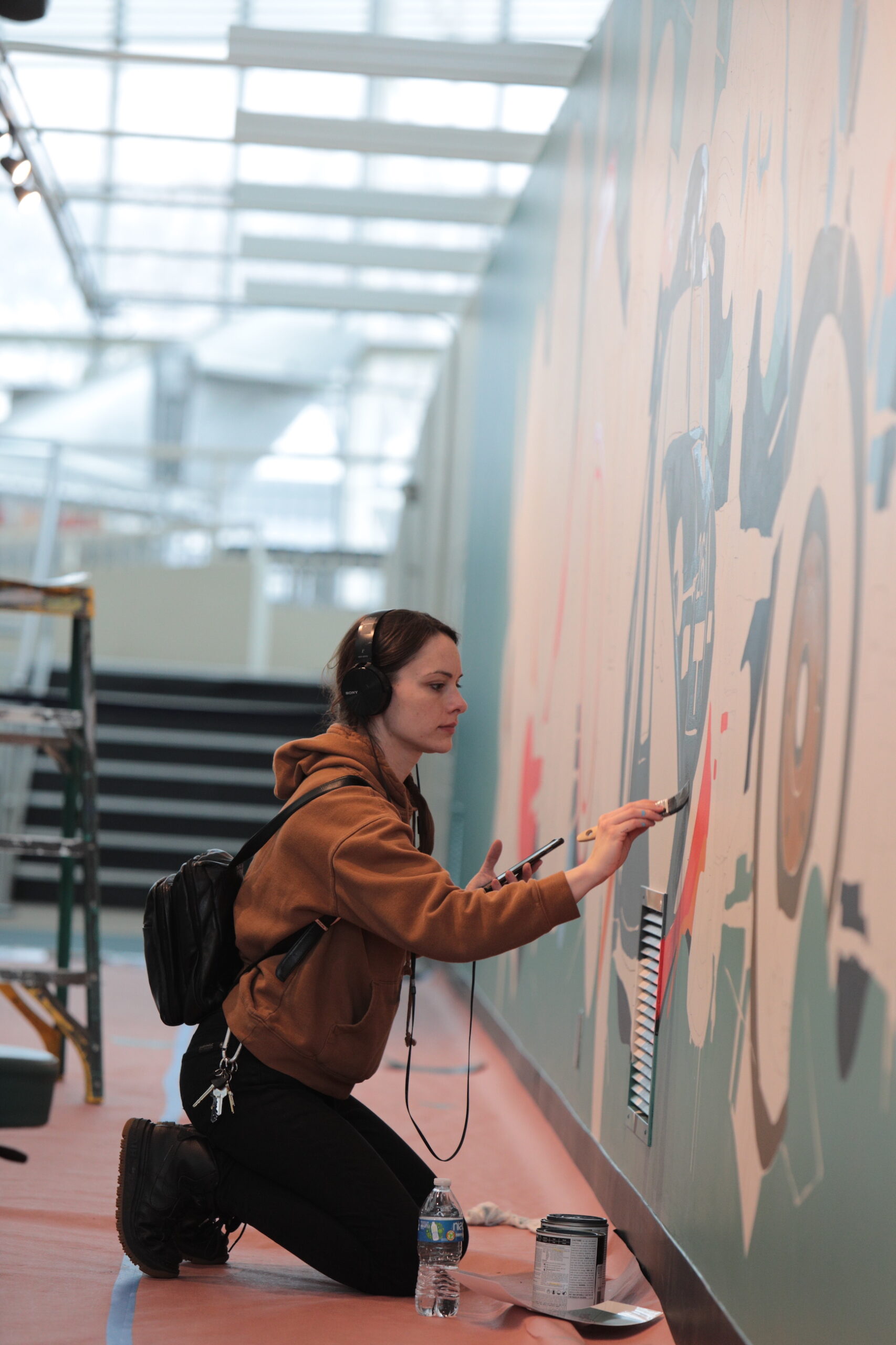 A woman with long brown hair wearing an orange sweatshirt paints a mural on a wall.