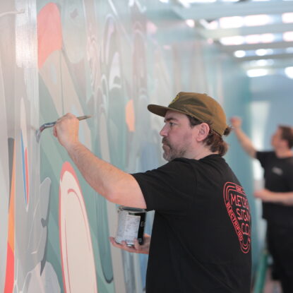 A white man wearing a baseball cap and black shirt paints a mural on a wall.