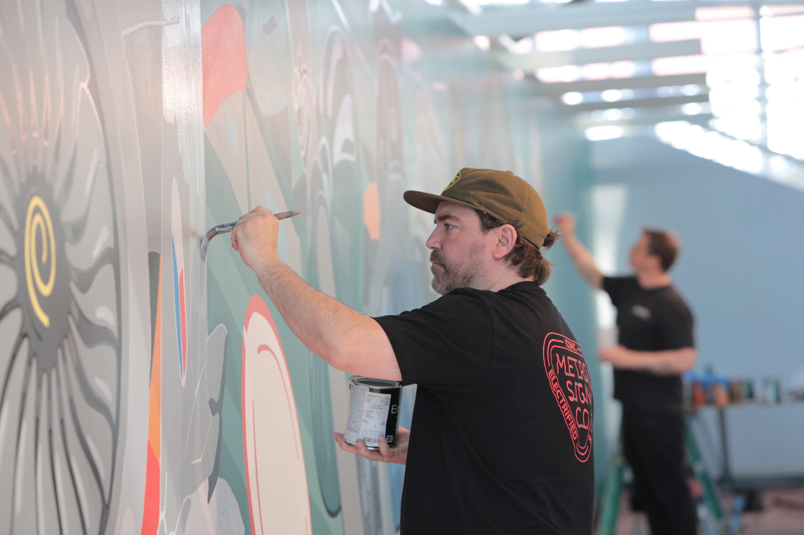 A white man wearing a baseball cap and black shirt paints a mural on a wall.
