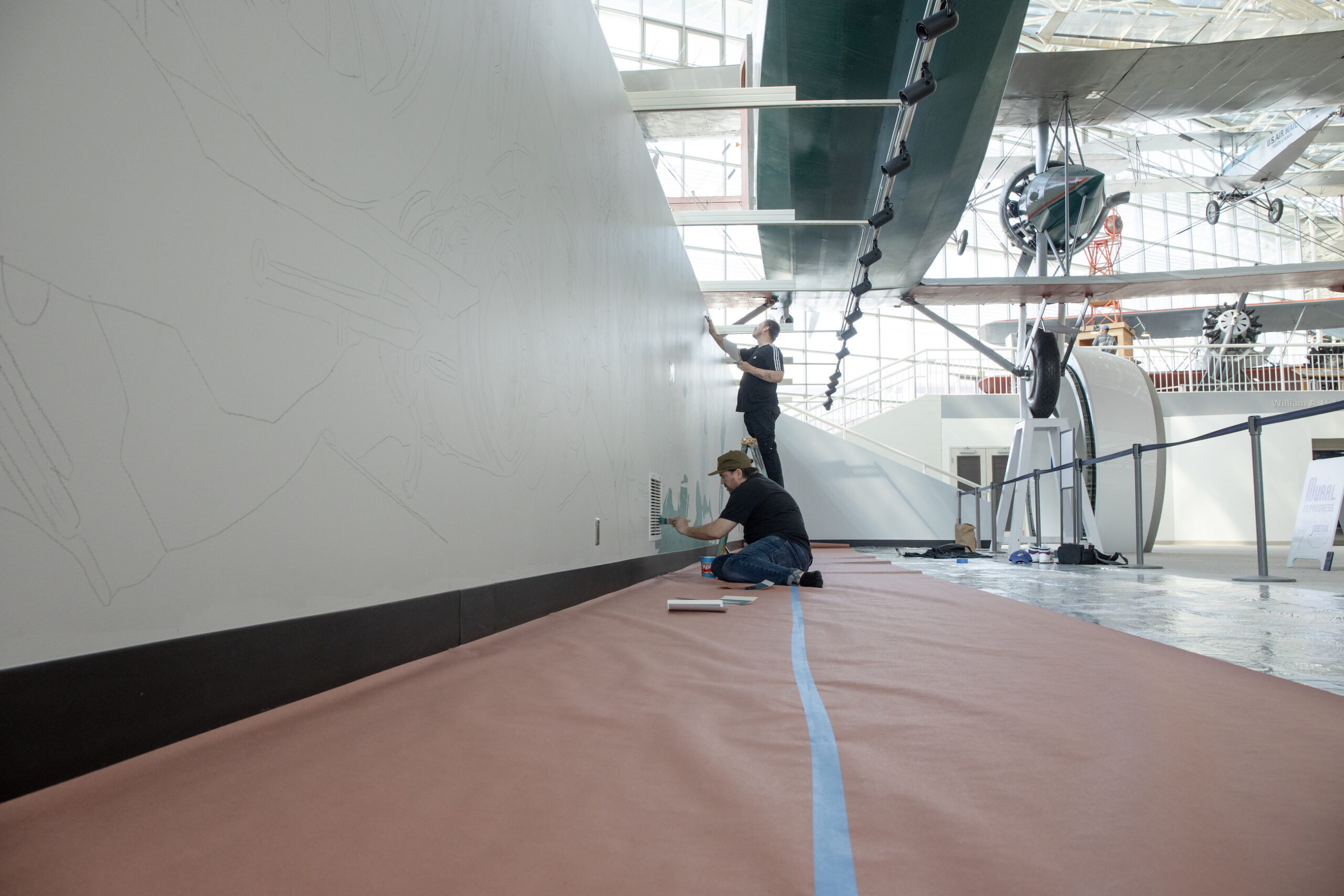 Artist Joe Nix sitting on the ground painting a mural in the Great Gallery with an assistant.
