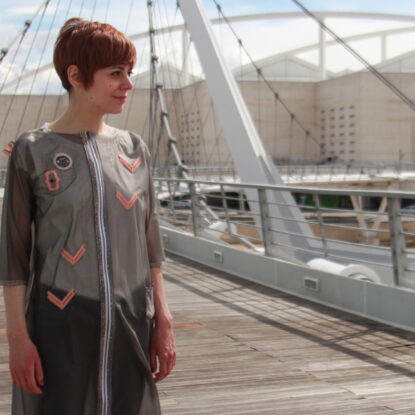 A person standing on a bridge wearing a shear grey garment. The garment resembles a raincoat adorned with copper elements and LEDs.