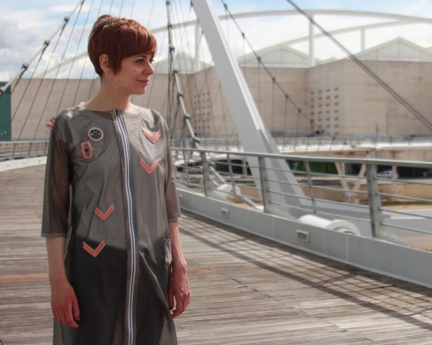 A person standing on a bridge wearing a shear grey garment. The garment resembles a raincoat adorned with copper elements and LEDs.