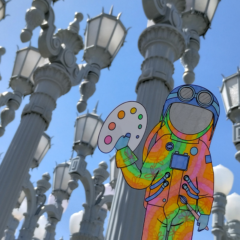 A colorfully decorated paper astronaut in the foreground with tall street lamps in the background.