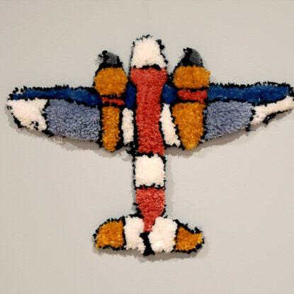 An artwork made of tufted orange, white and blue yarn in the shape of a plane hanging on a wall.