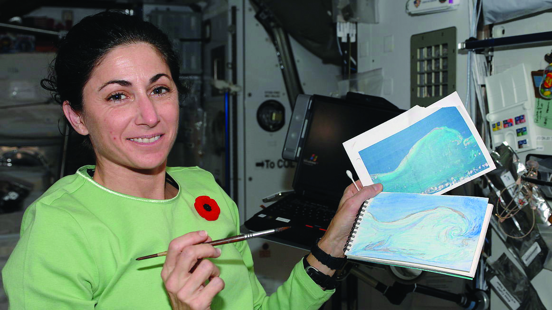 A woman with dark hair and bright green sweatshirt shows off watercolor paintings in the International Space Station