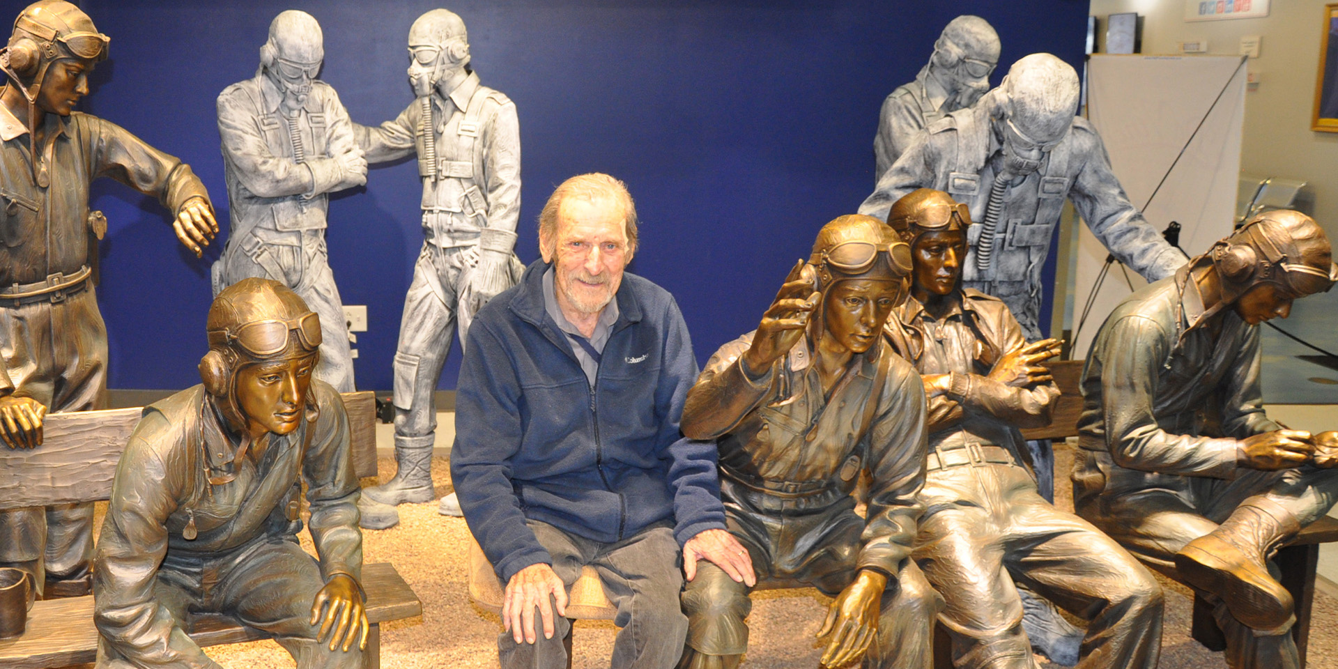 A white man with white hair sits among bronze statues featuring WWII serviceman.