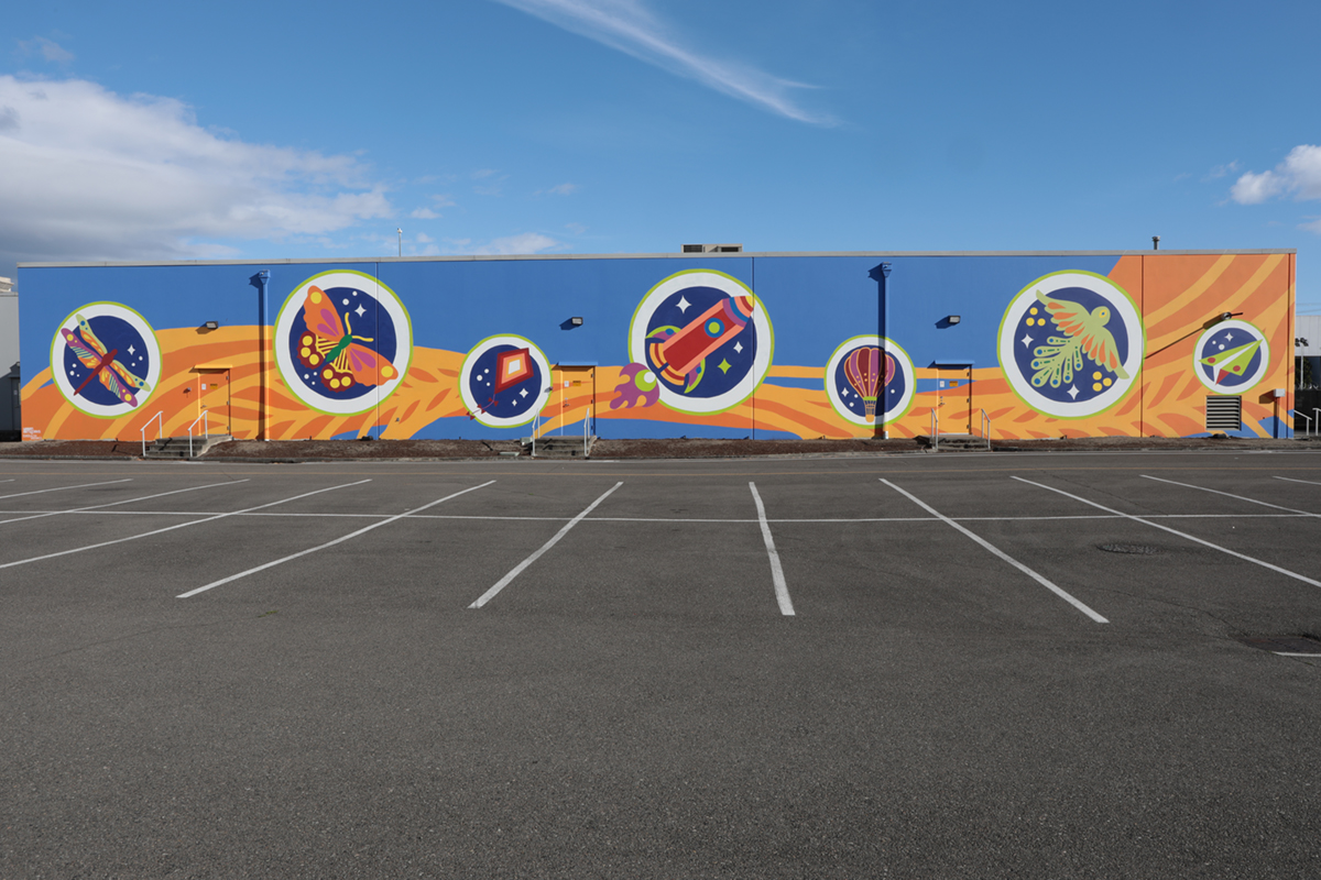 A wide, full view of a brightly painted wall mural of purples, yellows and oranges with a large, empty parking lot visible in front