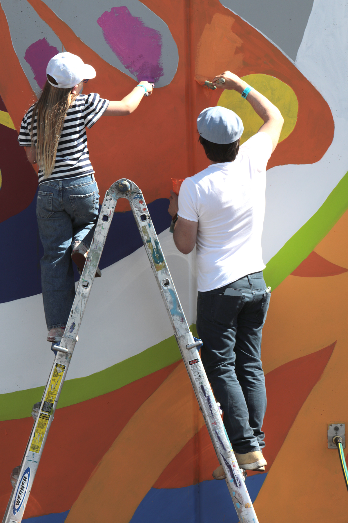 A young child and parent stand on a ladder painting a wall mural.