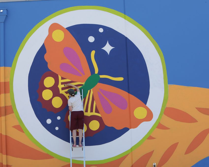 A person stands atop a ladder panting a large butterfly on a wall